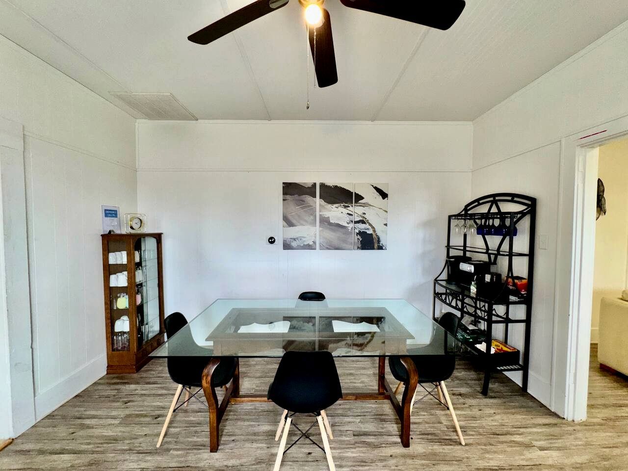 A dining area with a glass table, black chairs, a ceiling fan, a wall-mounted picture, a wooden cabinet, and a black shelving unit.