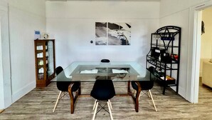 Un coin repas avec une table en verre, des chaises noires, un ventilateur de plafond, un tableau accroché au mur, un meuble en bois et une étagère noire.