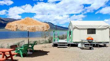 A beachside RV setup with a thatched umbrella, green chairs, a red picnic table, and a white RV with an orange gas tank.