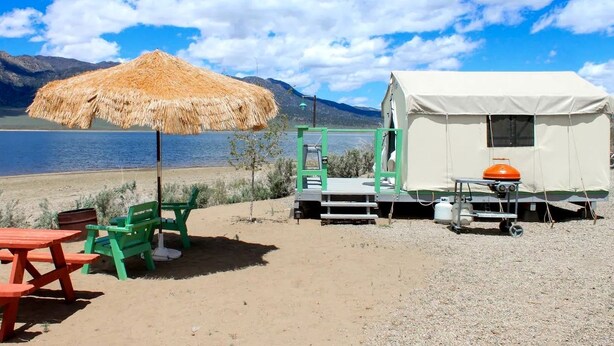 Un camping junto a la playa con una sombrilla de paja, sillas verdes, una mesa de picnic roja y una caravana blanca con tanque de gas naranja.