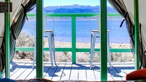 Una carpa con vistas a un lago y montañas, una cama con colcha y una silla afuera.