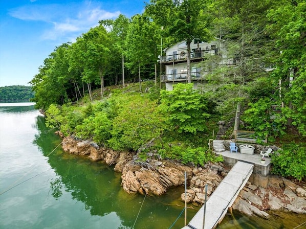 A lakeside cabin with a dock, surrounded by dense forest and a clear view of the water.
