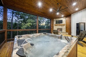 A hot tub on a wooden deck with a stone fireplace and a television. - Secluded Luxury Cabin for Couples with Soaker Tub & Forest Views in Oklahoma (Broken Bow)