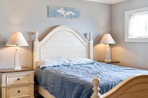 A bed with a white headboard, two bedside tables with lamps, a blue bedspread, and a wall with a 'Life is better up north' sign.