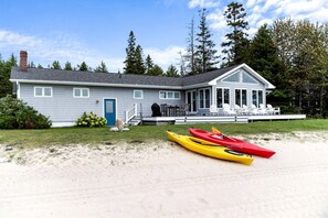Una casa con porche cubierto, una puerta azul y dos kayaks en la arena.