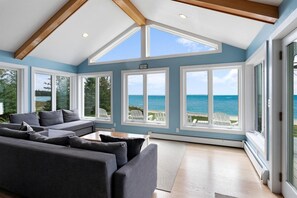 A spacious living room with a large grey sofa, multiple windows offering ocean views, and a wooden ceiling structure.