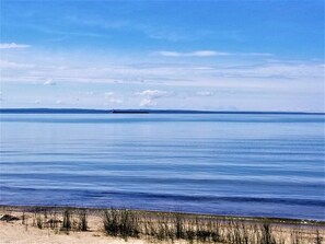 A calm body of water with a distant ship, clear sky, and sandy beach with sparse vegetation.