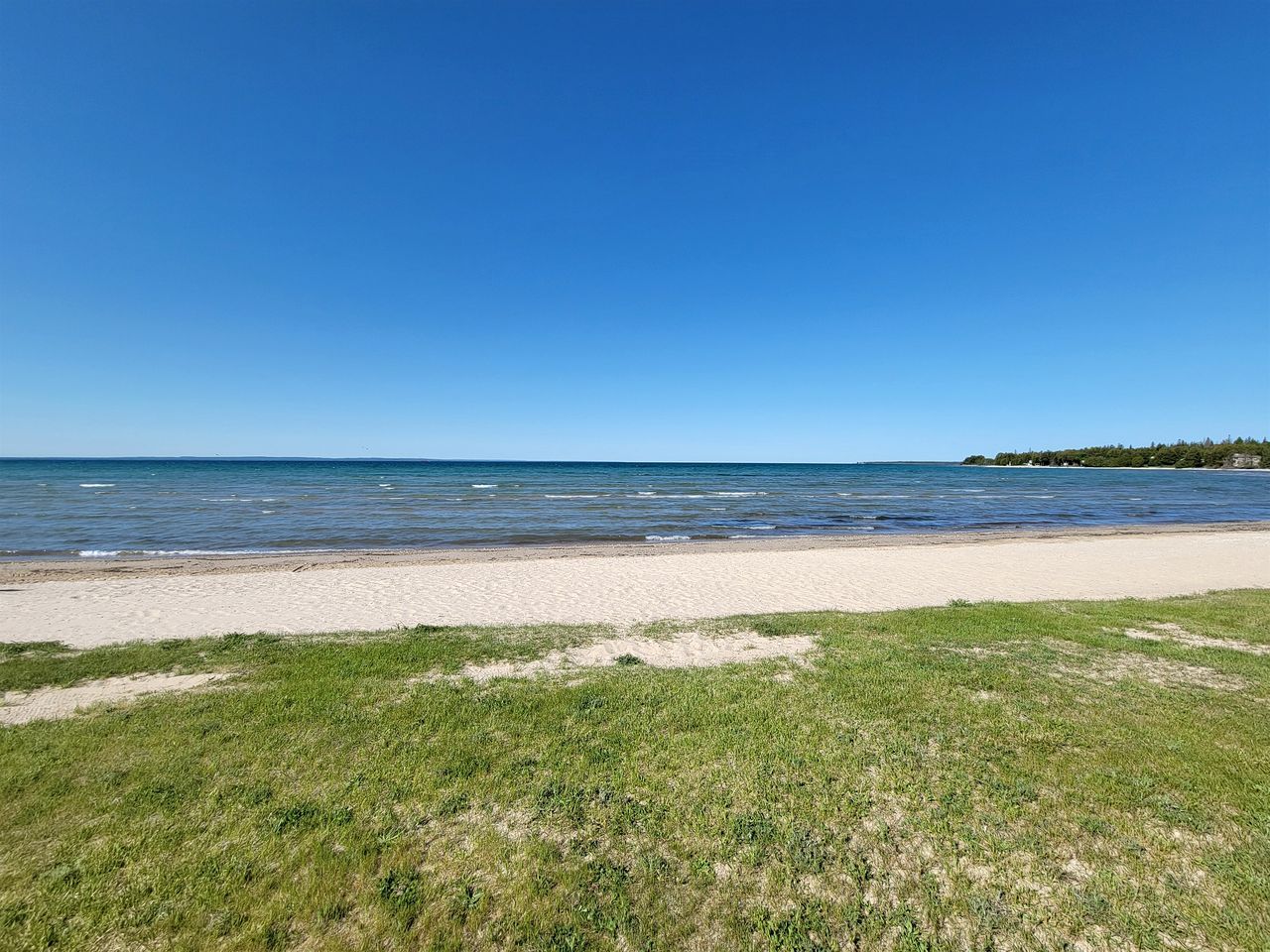 Une plage avec une eau d’un bleu limpide, un rivage sablonneux et une zone herbeuse.