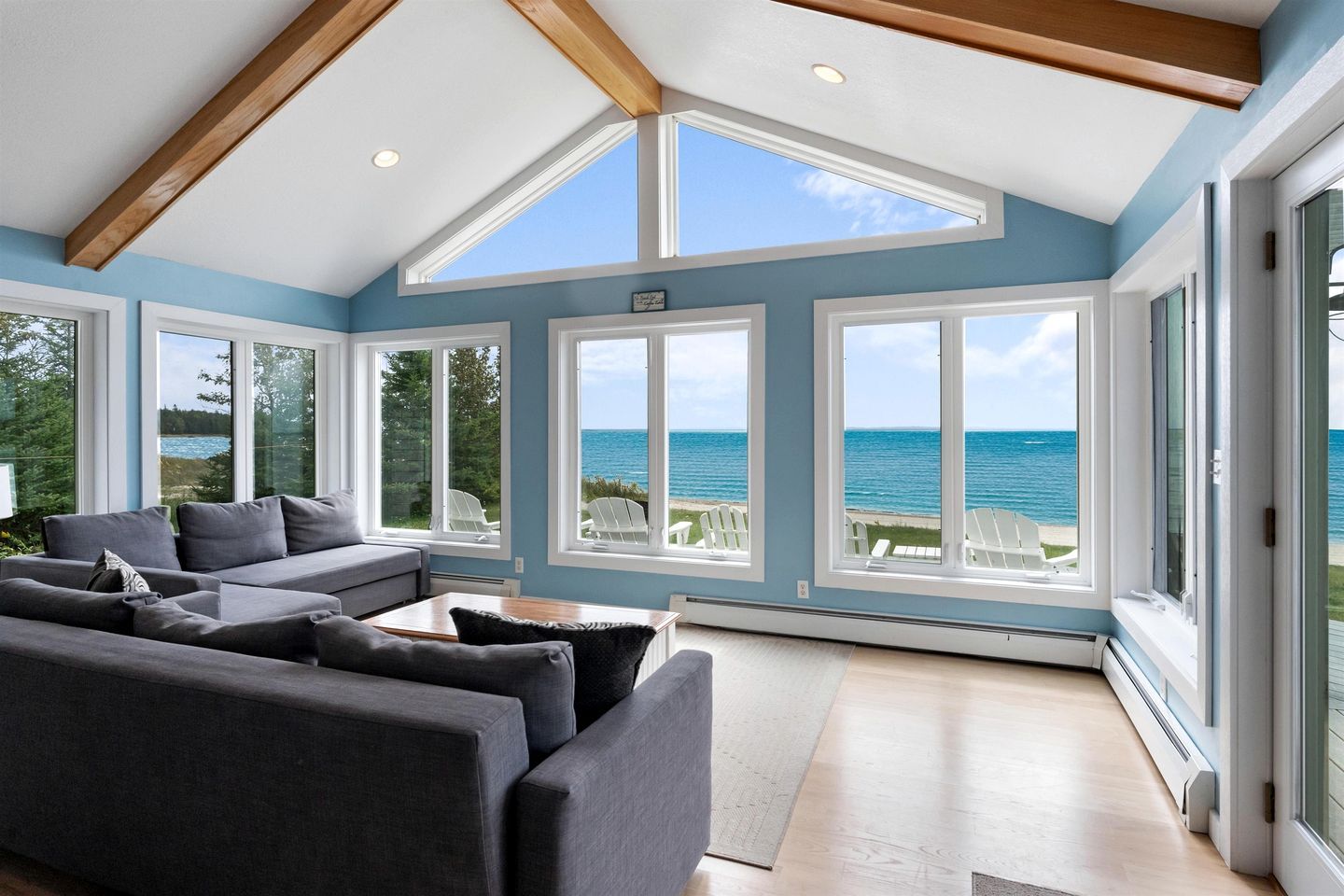 A spacious living room with a large grey sofa, multiple windows offering ocean views, and a wooden ceiling structure.
