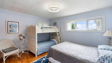 Een slaapkamer met stapelbed, raam, stoel, ventilator en een ingelijste foto aan de muur.