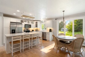 Una cocina moderna con muebles blancos, electrodomésticos de acero inoxidable, una isla grande con taburetes y un comedor con sillas de mimbre.