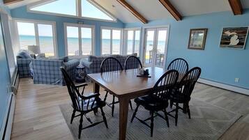 Un comedor con mesa y sillas de madera, ventilador de techo y grandes ventanas con vista al océano.