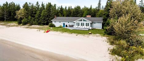 A beachfront property with a large sandy beach, a white house with a porch, and dense forest in the background.