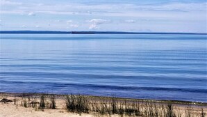 A calm body of water with a distant ship, clear sky, and sandy beach with sparse vegetation. - Beautiful Lakefront Cottage with Fire Pit in St. Ignace, Michigan (St. Ignace)