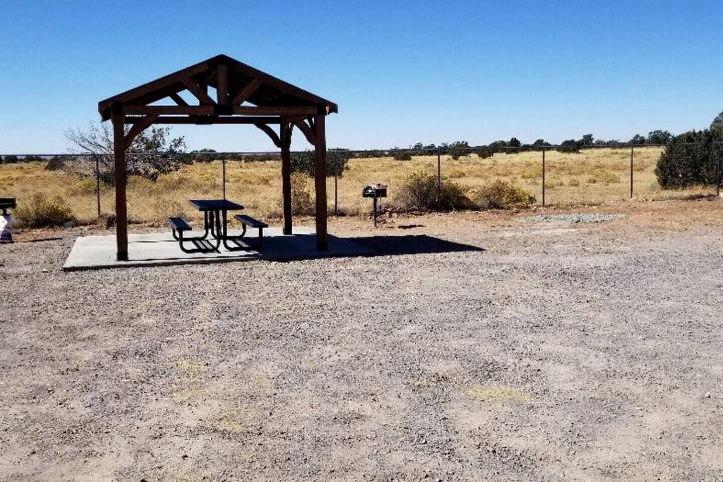 A wooden gazebo with picnic tables and a mailbox in a dry, open field.