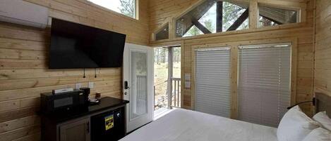 Ein Zimmer in einer Holzhütte mit einem Bett, einem Flachbildfernseher, einem Schreibtisch mit einem Computer und einer Schiebetür nach draußen.