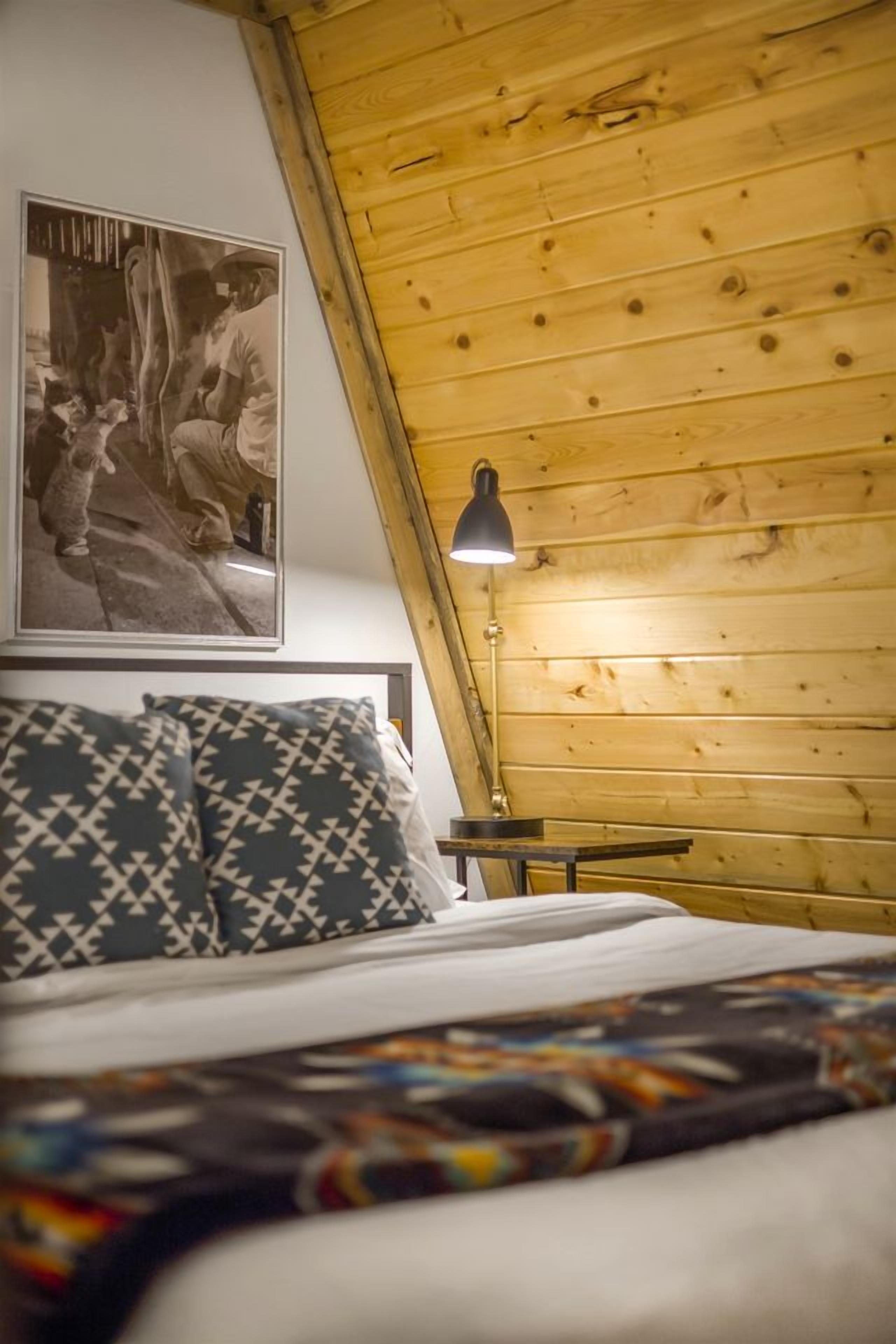 Une chambre avec un plafond en pente en bois, un lit agrémenté d’oreillers à motifs, une table de chevet avec une lampe et un tableau encadré au mur.