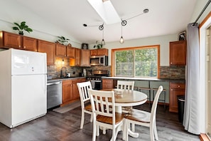 A kitchen with wooden cabinets, a white refrigerator, a dining table with chairs, and a window with a view of trees.