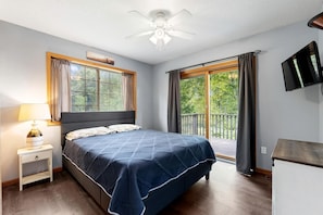 A bedroom with a bed, a nightstand, a ceiling fan, a window with curtains, and a TV mounted on the wall.