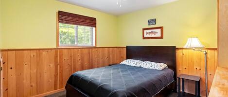 Quarto com cama, janela com persianas, ventilador de teto, abajur e quadro na parede.