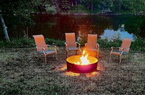 A campfire with chairs around it, set in a grassy area near a body of water. - Riverfront Cabin Incredible for Outdoor Enthusiasts with Kayaks in Hessel, Michigan (Hessel)