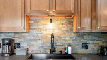 A kitchen with wooden cabinets, a stone backsplash, a stainless steel sink, a coffee maker, and a soap dispenser.