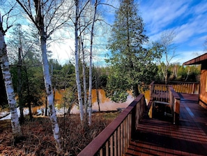 A wooden deck with a metal railing overlooking a forested area and a body of water. - Riverfront Cabin Incredible for Outdoor Enthusiasts with Kayaks in Hessel, Michigan (Hessel)