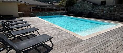 A swimming pool with lounge chairs, a wooden deck, and stone buildings in the background.