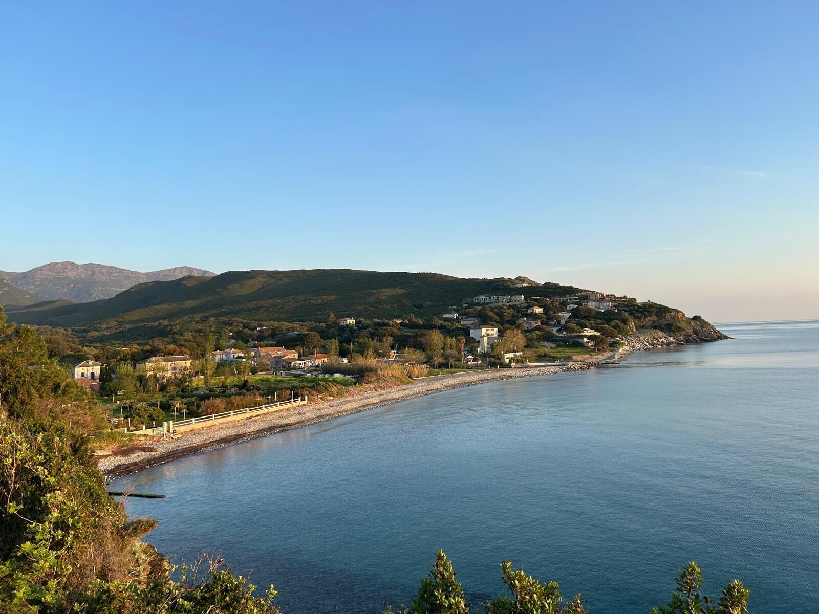 Une ville côtière avec des maisons qui bordent le rivage, un ciel dégagé et des montagnes en arrière-plan.