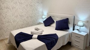 Et hotelværelse med væg i marmorlook, en hvid seng med marineblå puder, et hvidt natbord med lampe, og et pænt foldet håndklæde på sengen.