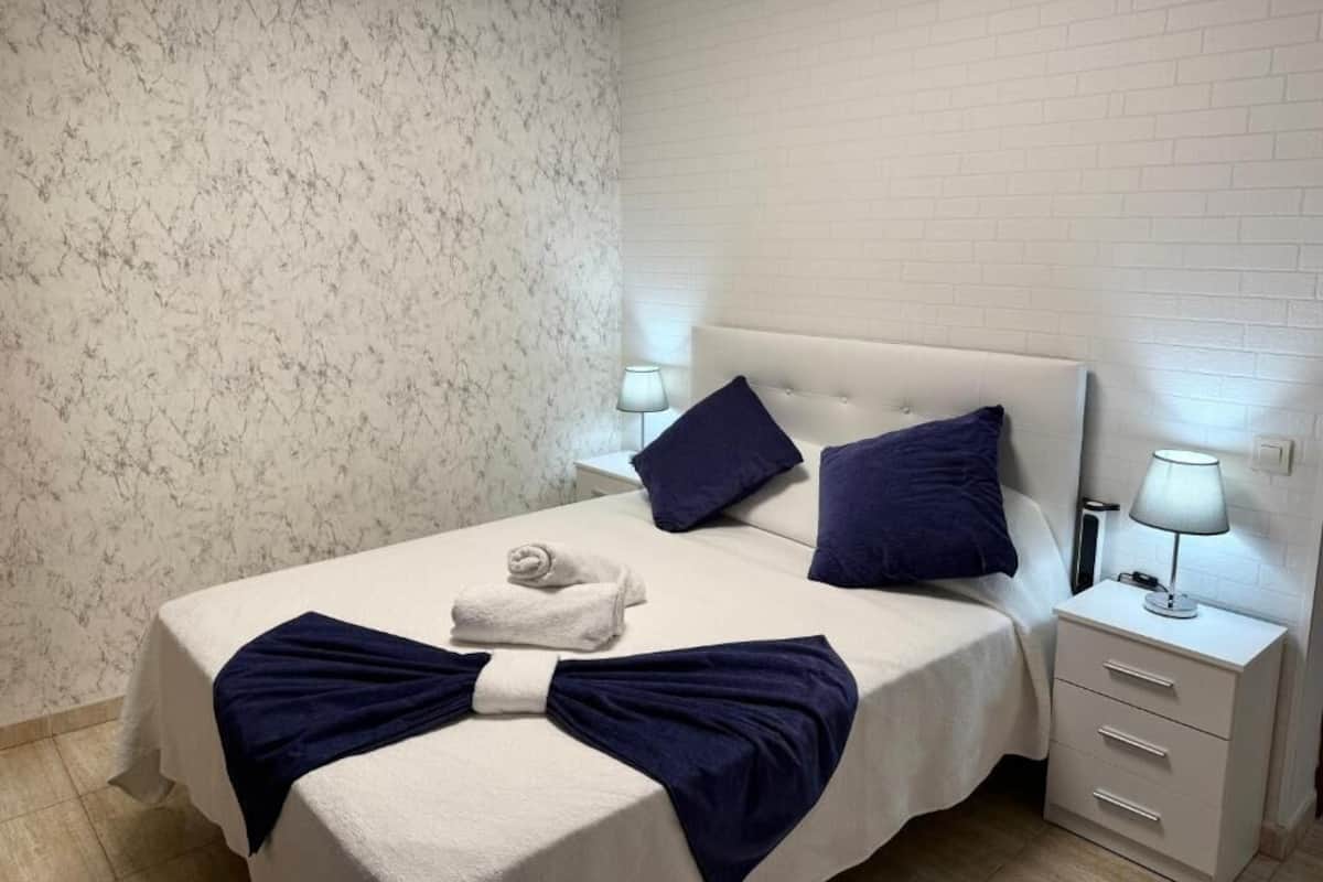 Habitación de hotel con pared de diseño de mármol, cama blanca con almohadas azul marino, mesita de noche blanca con lámpara y una toalla doblada cuidadosamente sobre la cama.