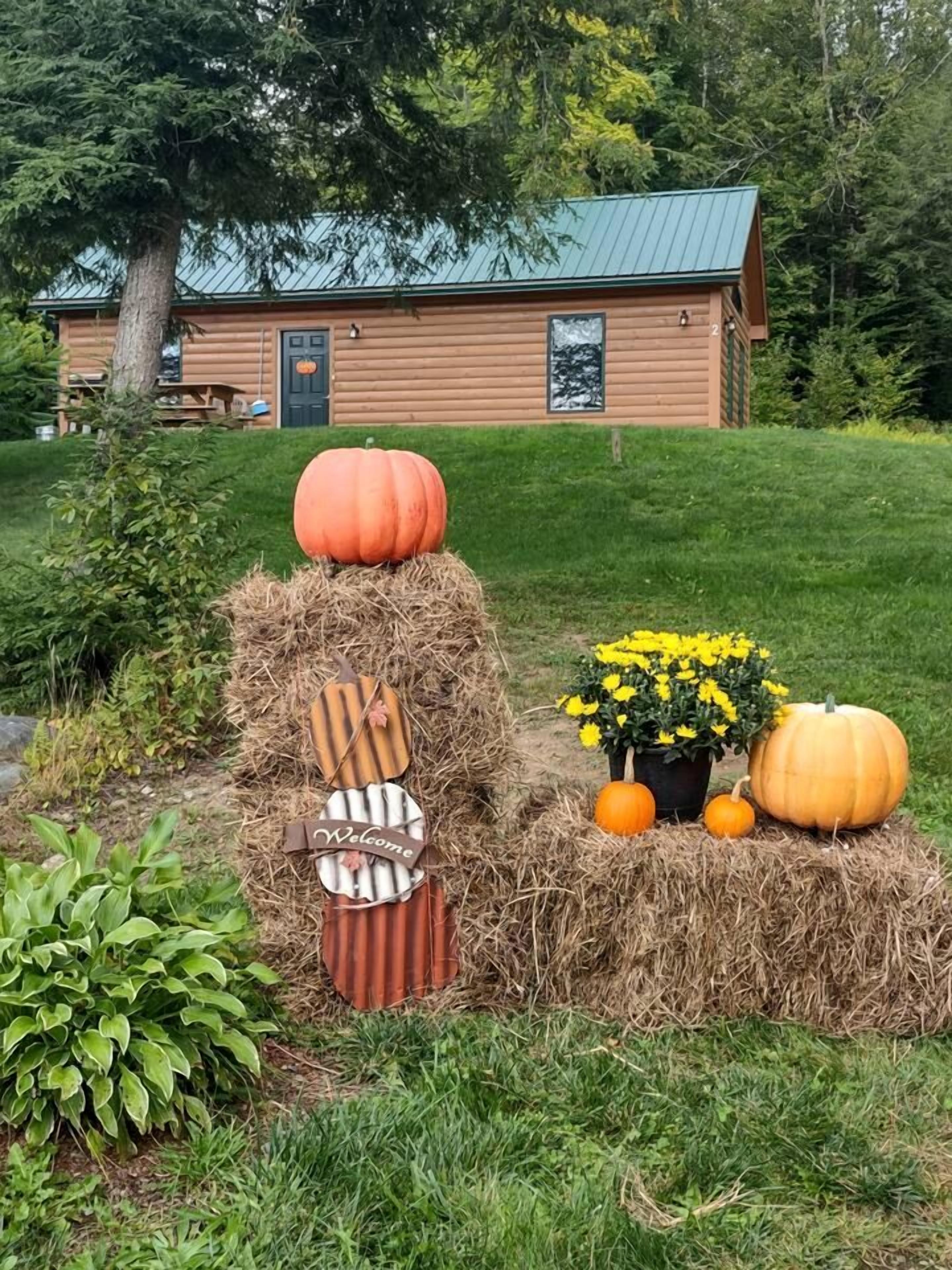 A rustic cabin with a green roof, surrounded by a well-maintained lawn and a hay bale decoration with pumpkins and a welcome sign.