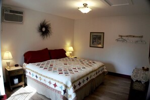 A bedroom with a bed, two bedside lamps, a wall-mounted air vent, a decorative wall piece, a framed picture, and a small table with a candle.