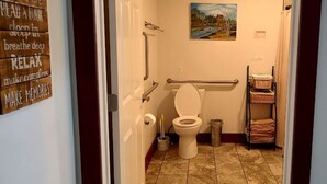 A bathroom with a toilet, handrails, a painting on the wall, and a shelf with towels. - Handicapped Accessible Log Cabin Rental near Smugglers' Notch Park, Vermont (Hyde Park)