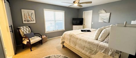 A bedroom with a bed, a chair, a desk, a ceiling fan, a TV, and artwork on the walls.