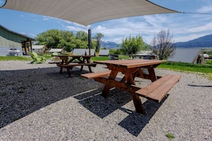 Outdoor picnic tables with a shaded area, surrounded by gravel, with buildings and trees in the background.
