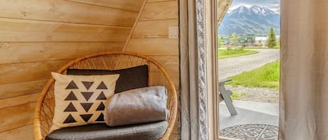 Un interior de madera con una silla de mimbre, cojines y vistas a la montaña a través de una puerta.