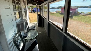 A covered porch with a table and chairs, large windows with a view of a lake and buildings, and an air conditioning unit. - Lakefront Cottage Rental near Bear Brook State Park in Northwood, New Hampshire (Northwood)