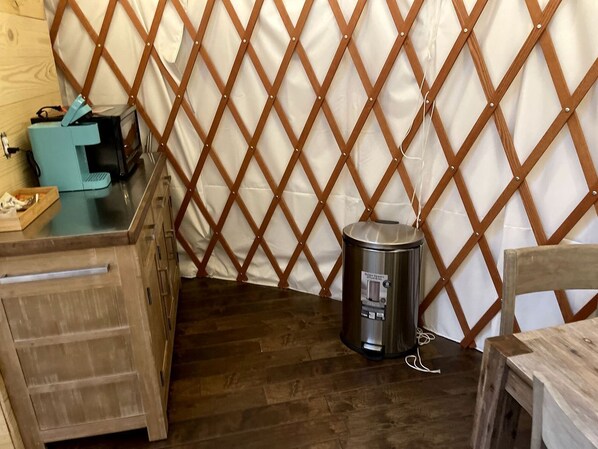 Una habitación con piso de madera, un bote de basura metálico, una mesa de madera y un cortinero de madera con un diseño de diamantes.