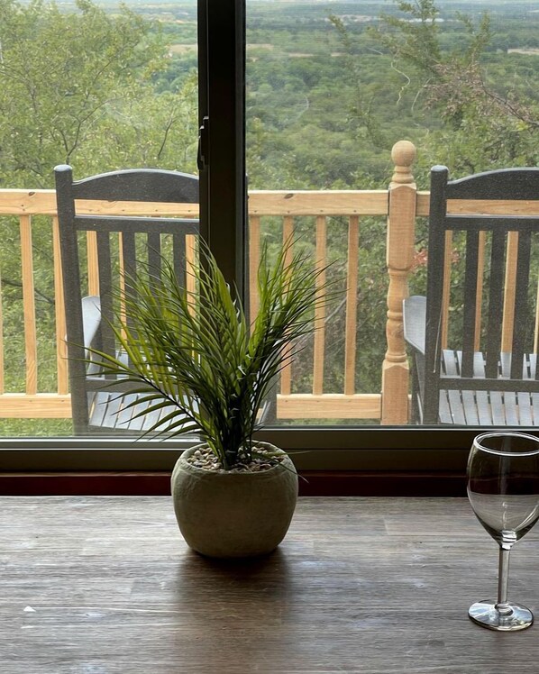 Una planta en maceta sobre una mesa de madera junto a una ventana con vista a un balcón y paisajes al aire libre.