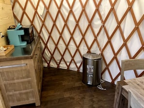 Ein Zimmer mit Holzboden, einem Metallmülleimer, einem Holztisch und einem weißen Vorhang mit einem rautenförmigen Holzrahmen.