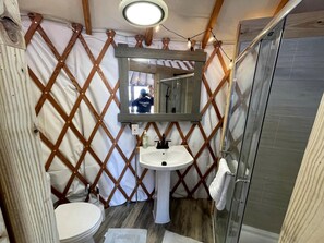 A compact bathroom with a wooden lattice partition, a pedestal sink, a mirror, and a glass shower enclosure.