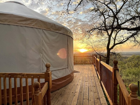 A wooden deck leading to a large, round, covered structure with a view of a sunset and trees.
