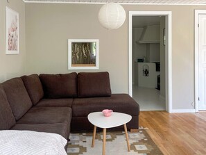 A living room with a brown sofa, a white table with a pink bowl, a framed picture on the wall, and a doorway leading to a kitchen.