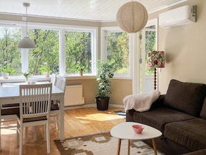 Une pièce comprenant une table à manger, des chaises, un canapé, une petite table avec un saladier, et de grandes fenêtres donnant sur un paysage verdoyant.