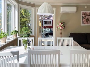Una habitación con una mesa y sillas blancas, una lámpara colgante, una ventana con plantas y un equipo de aire acondicionado.