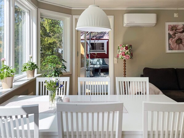A room with a white table and chairs, a hanging lamp, a window with plants, and an air conditioning unit.