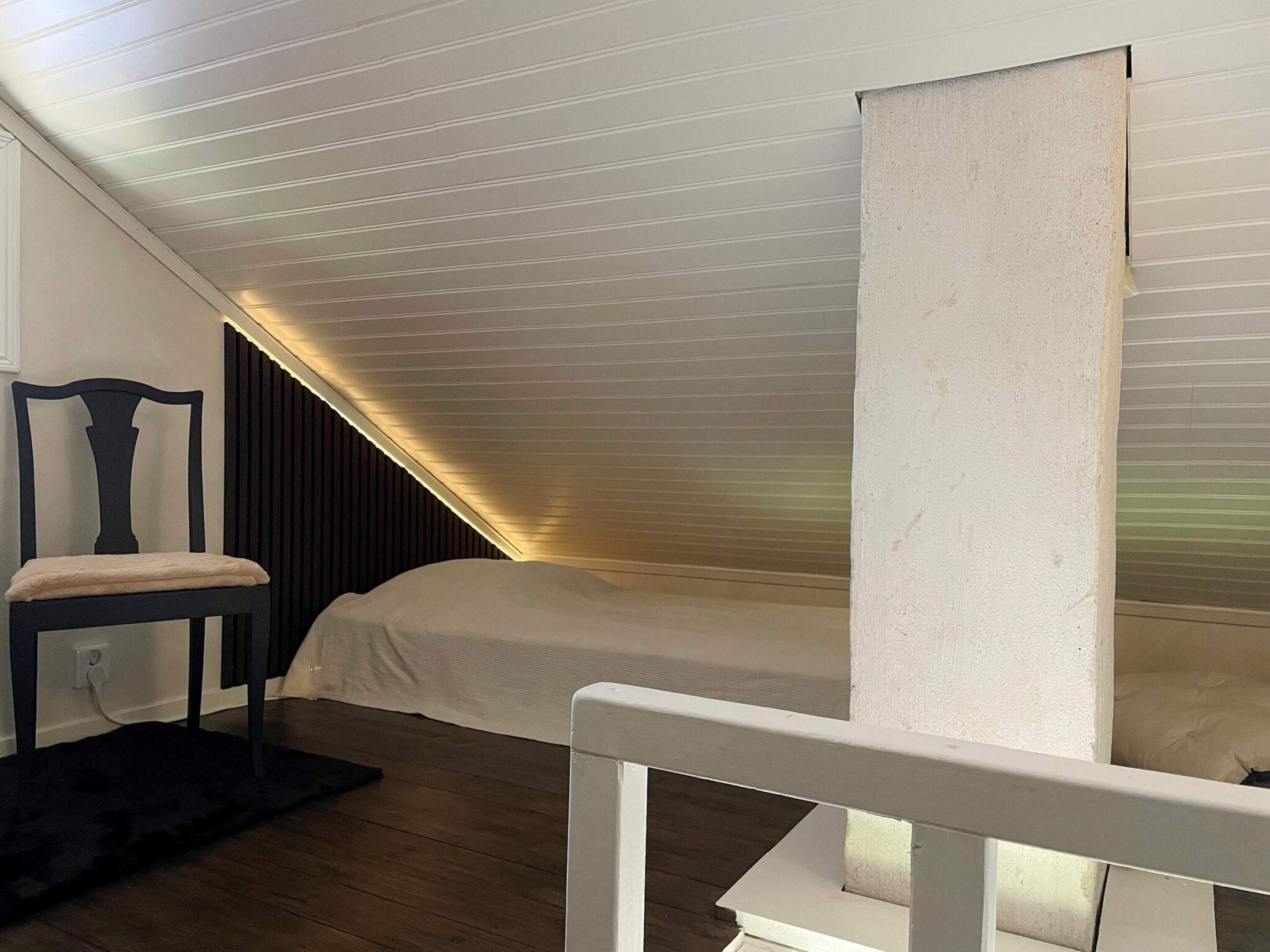 A room with a sloped ceiling, a bed, a chair, and a dark floor.