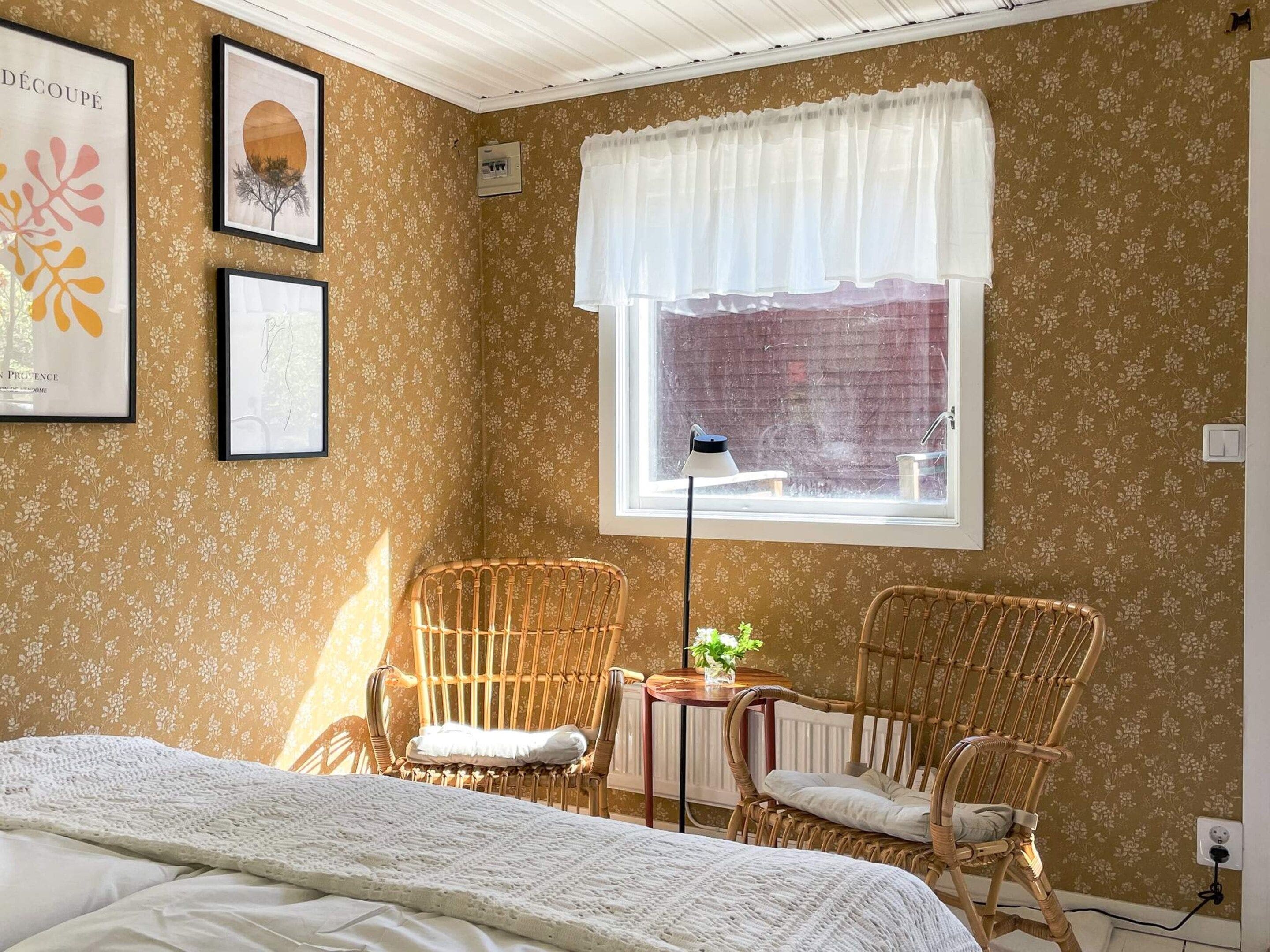 A room with a bed, two wicker chairs, a small table with a plant, a window with a white curtain, and floral wallpaper.