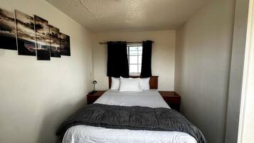 A single bed with a gray blanket, white pillows, and a wooden headboard. There is a window with black curtains, a bedside table with a lamp, and a wall-mounted picture frame.